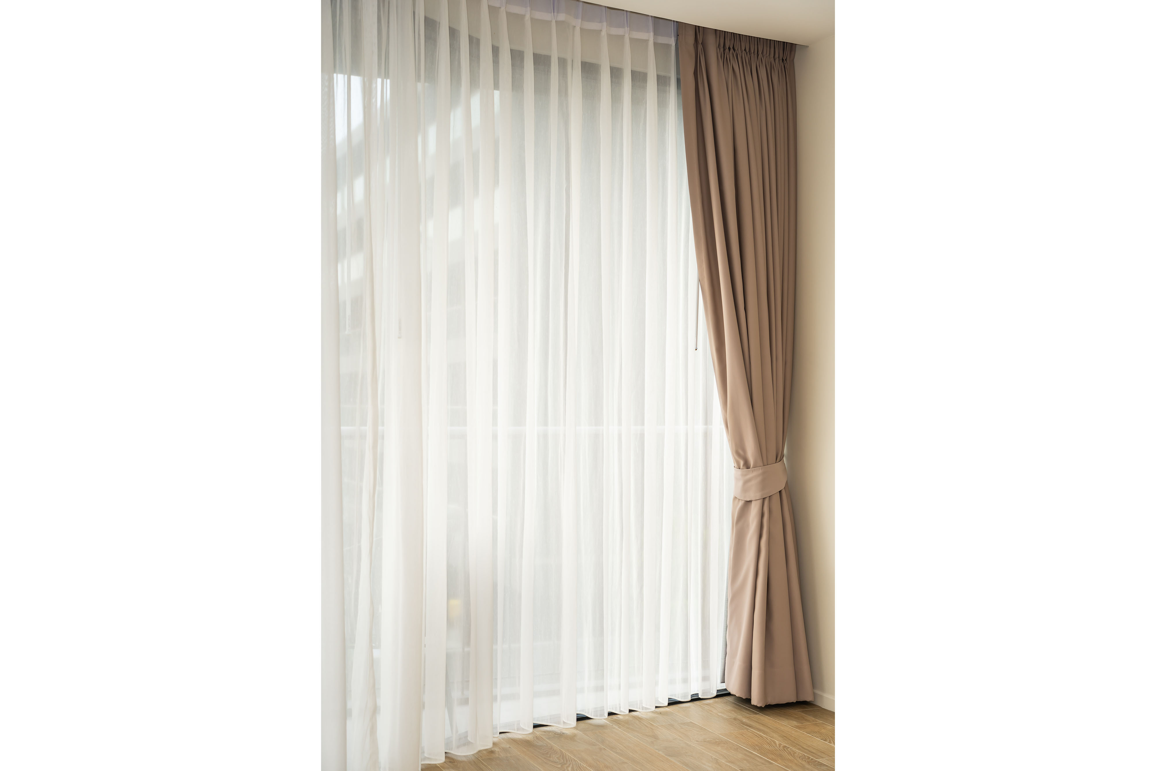 A floor-to-ceiling window layered with a sheer white voile panel beneath a heavier taupe drape, the outer curtain gathered back with a matching tieback.
