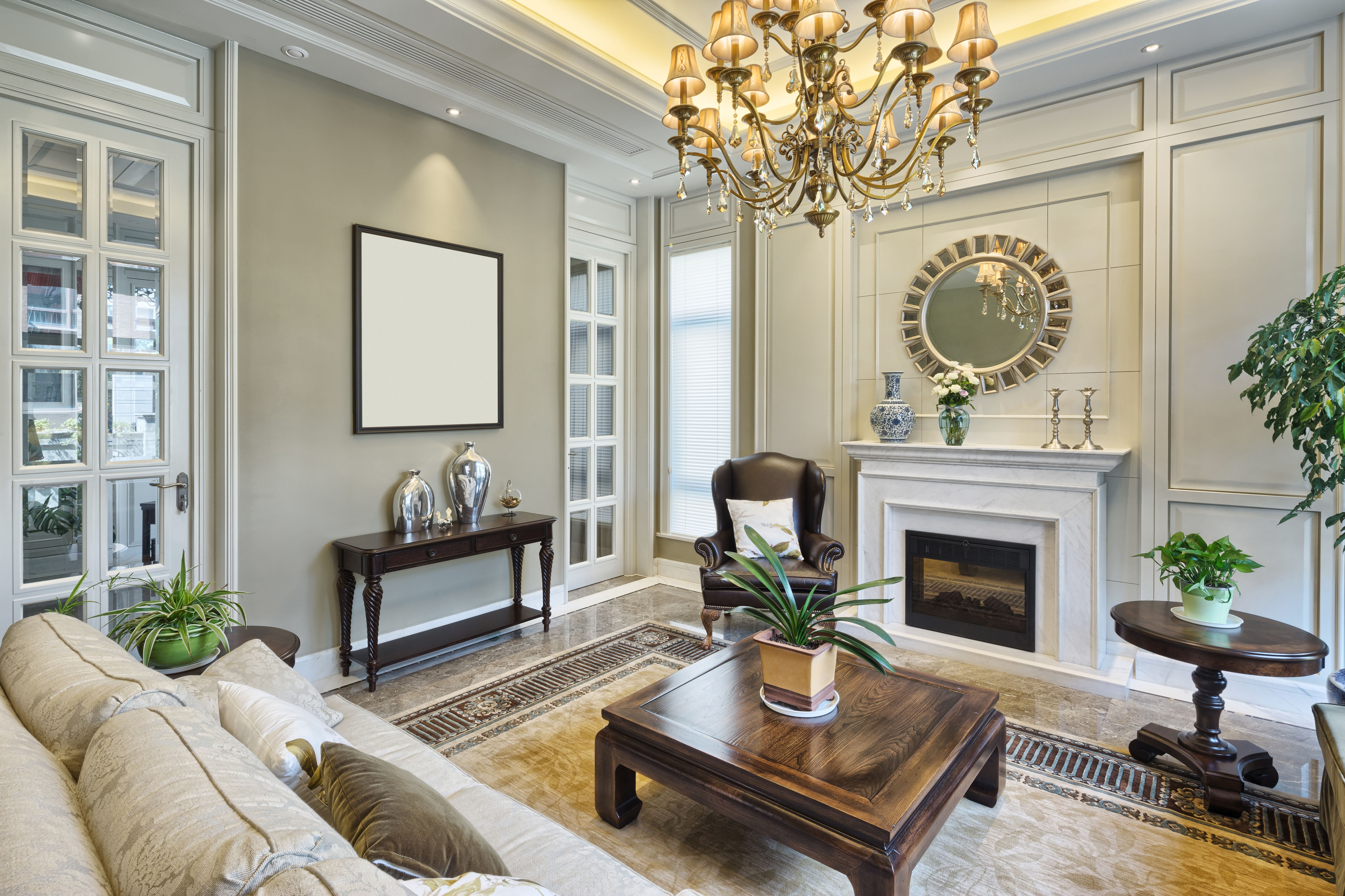A traditional living room with crown molding, a marble fireplace, and a dark wood console table topped with a mirror beneath a crystal chandelier.
