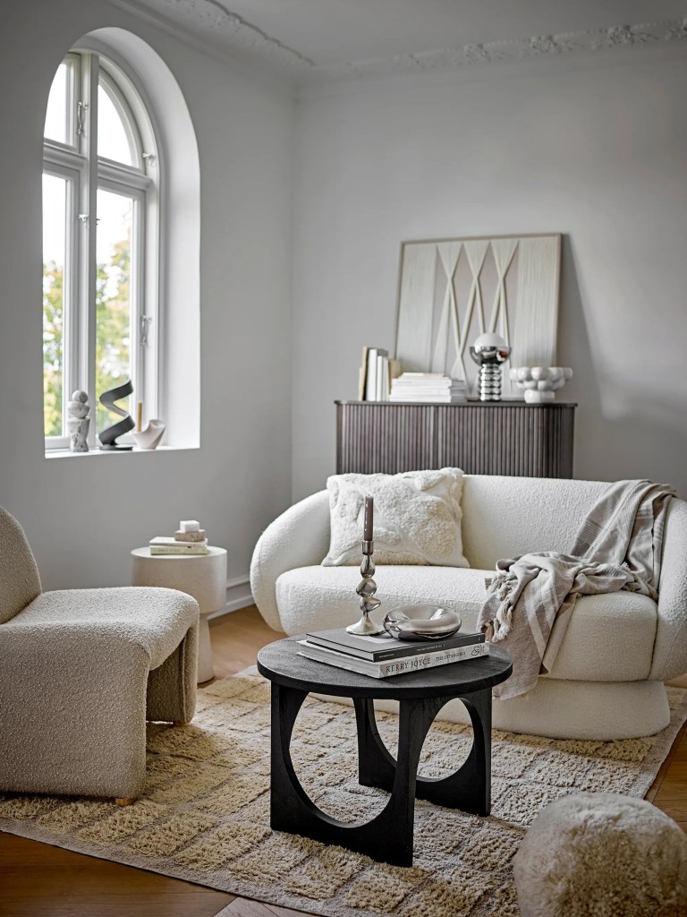 A cream sofa and two boucle swivel chairs arranged around a sculptural black coffee table beneath an arched window, grounded on a layered patterned rug.