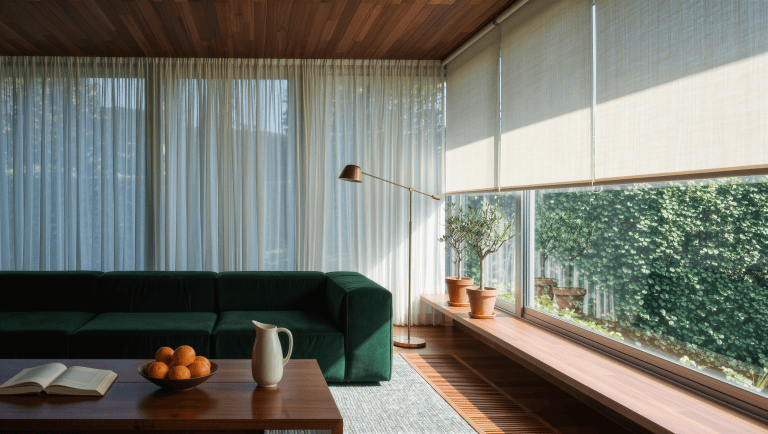 A living room with a floor-to-ceiling window wall softened by sheers and roller shades, the green sofa angled away from the glare.