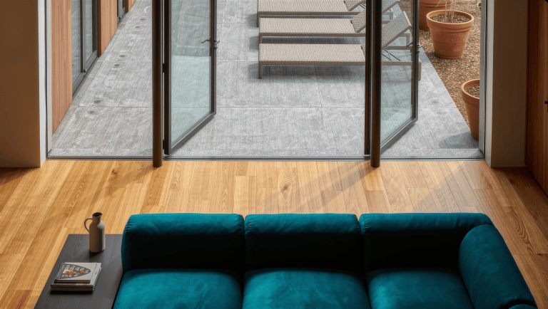 A living room with open steel-framed glass doors onto a patio, the teal sofa inside and woven lounge chairs outside sharing one sightline.