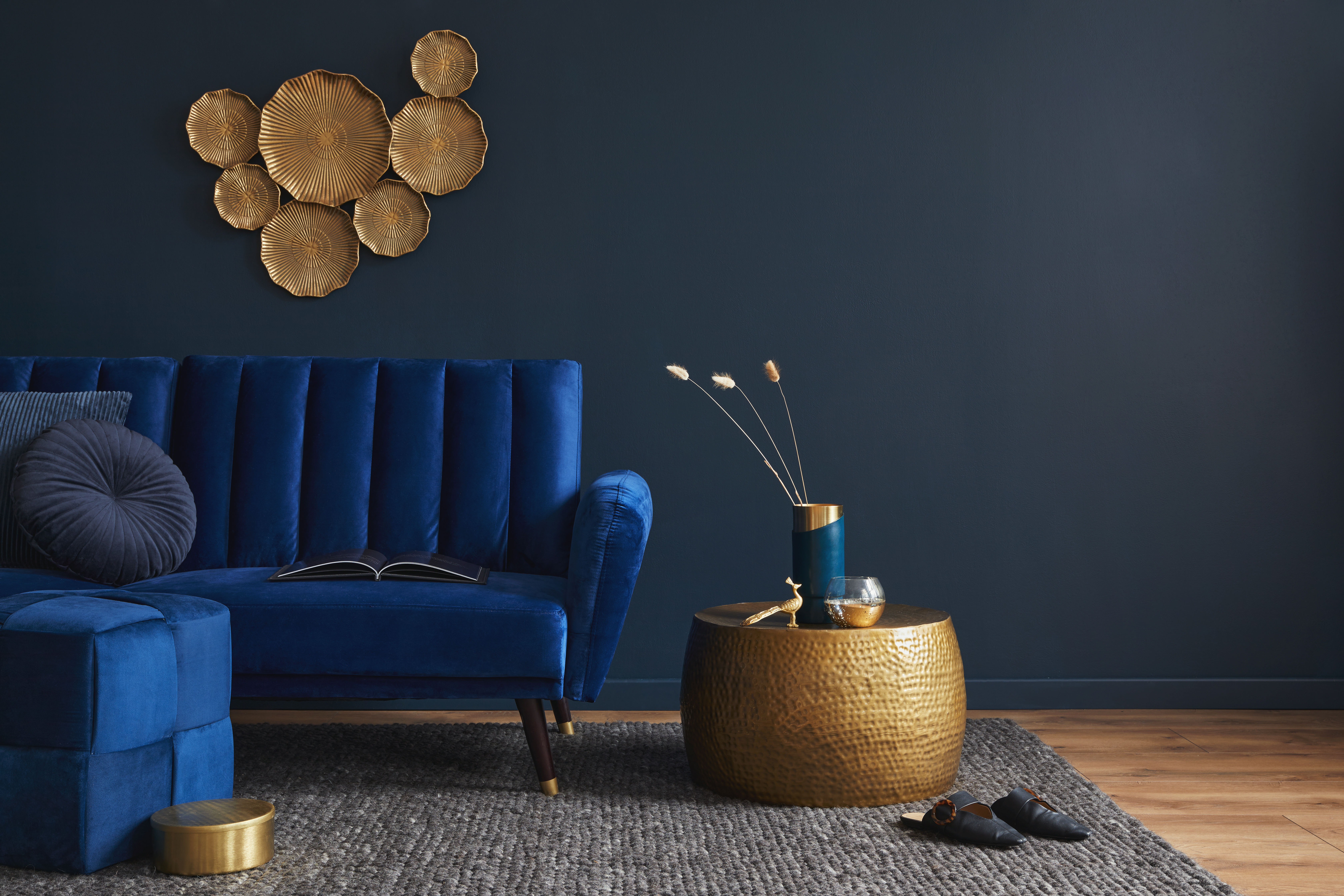A royal-blue channel-tufted velvet sofa with brass legs beneath a gold disc wall sculpture, paired with a hammered brass coffee table against a charcoal-navy wall.
