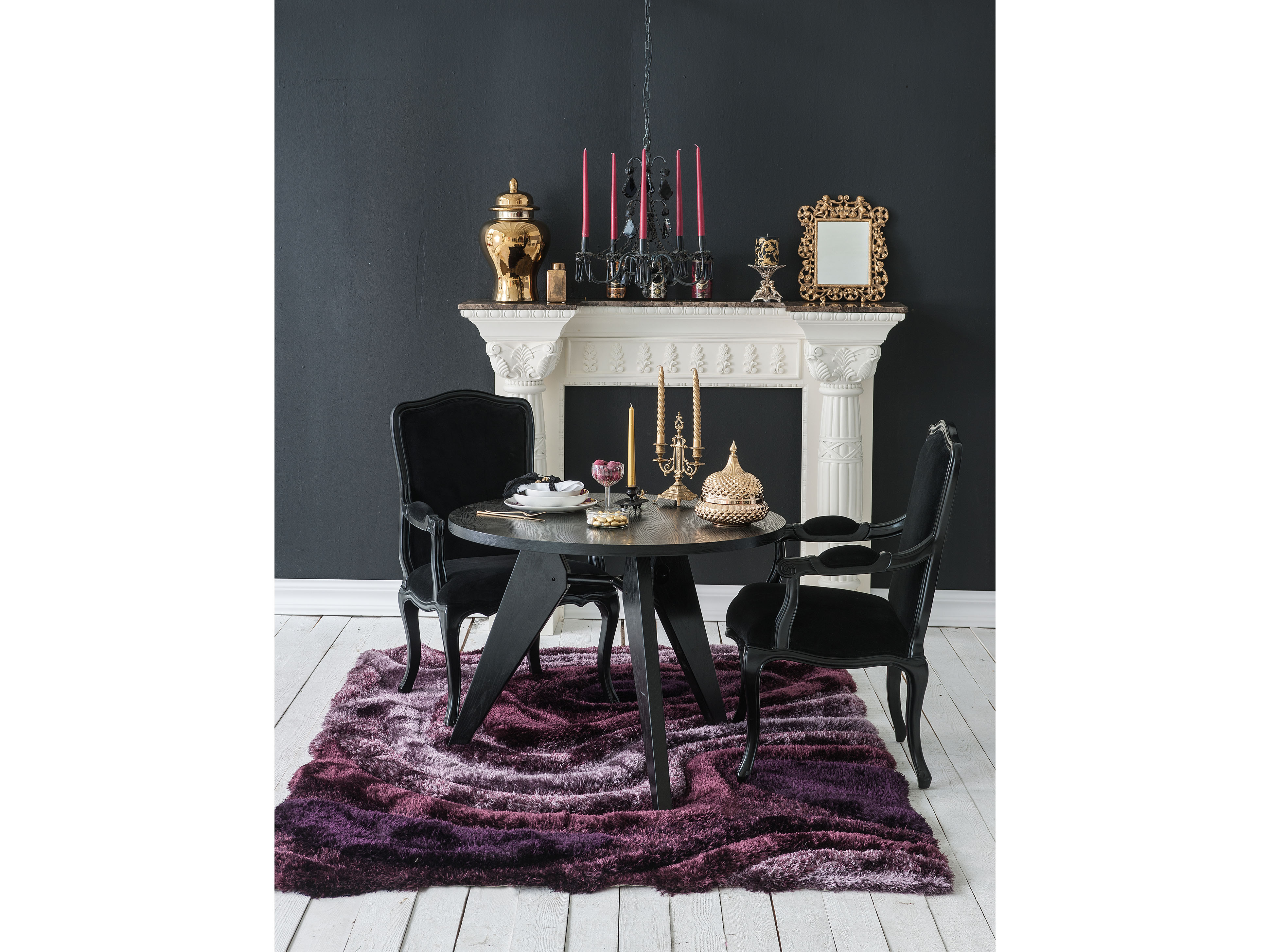 A dramatic black-walled dining room with a white marble mantel styled with brass candlesticks and gilt-framed art, anchored by a round black table and a plum shag rug.