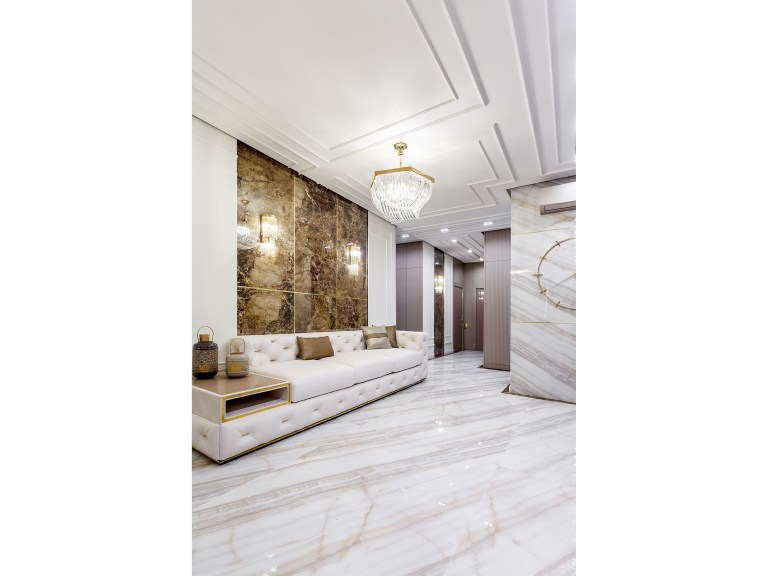 A glossy marble floor with veined stone banding sweeps through an open luxury living room, marking a clear route past a cream tufted sofa toward a gilded chandelier and a bronze mosaic accent wall.