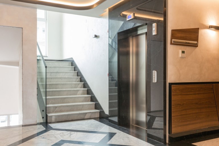 A polished marble-floored hotel corridor with a glass elevator leads past a wood-paneled wall and staircase, where warm wood tones meet the cool stone flooring.