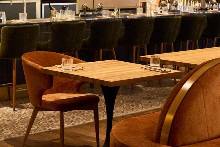 A curved emerald velvet banquette lines a warm dining nook, with wood tables on slender brass-toned legs and low pendant lighting overhead.