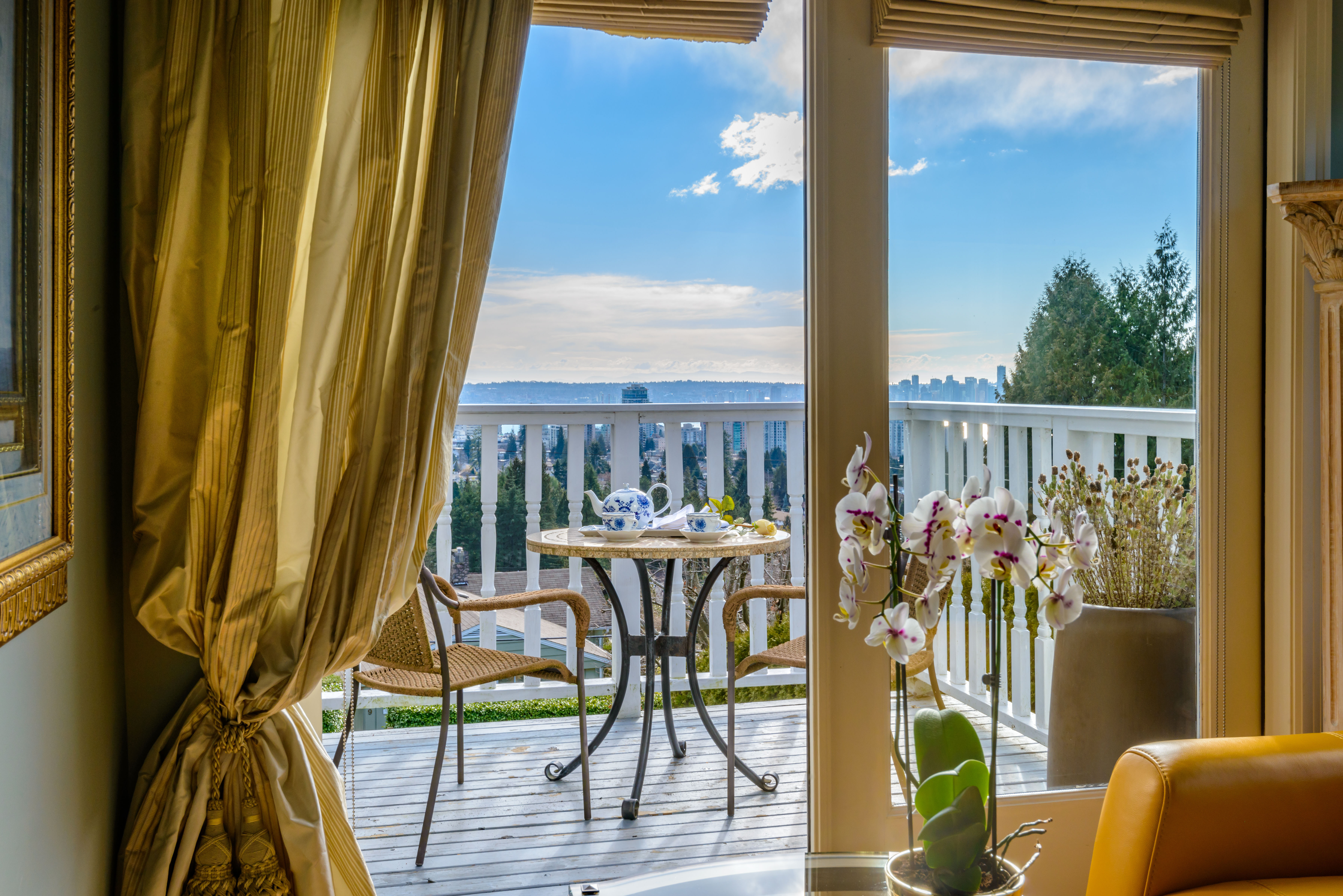 A small round bistro table dressed with orchids sits on a private balcony framed by gold drapery, opening onto a sea view.
