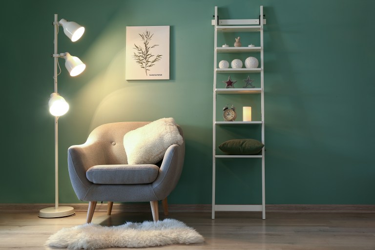 A tufted grey-blue velvet armchair sits against a deep emerald wall between a warm three-globe floor lamp and a ladder bookshelf styled with books, art, and a botanical print.