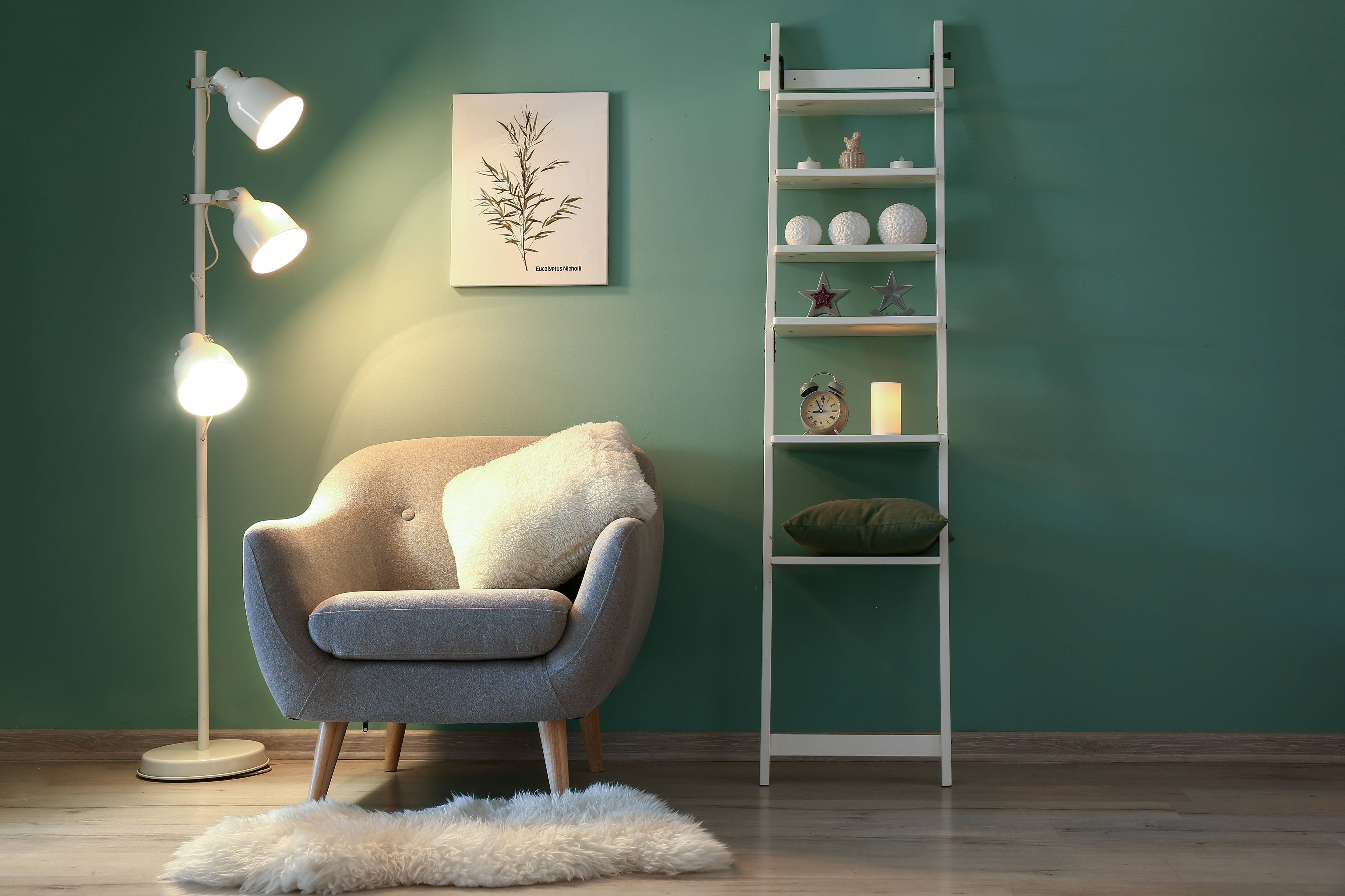 A tufted gray-blue velvet armchair sits against a deep emerald wall between a warm three-globe floor lamp and a ladder bookshelf styled with books, art, and a botanical print.
