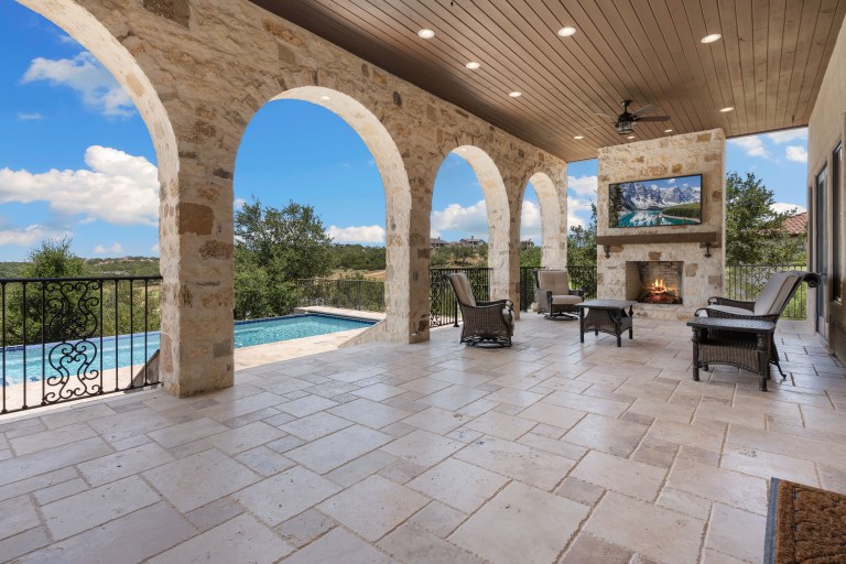 A stone-arched covered patio with a travertine floor, lounge seating and a stone outdoor fireplace set off to one side, framing a pool and hill-country view beyond.