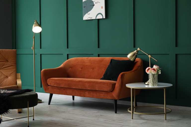 A burnt-orange velvet sofa sits against an emerald green paneled wall, with a brass floor lamp and a round marble side table styled with fresh flowers.
