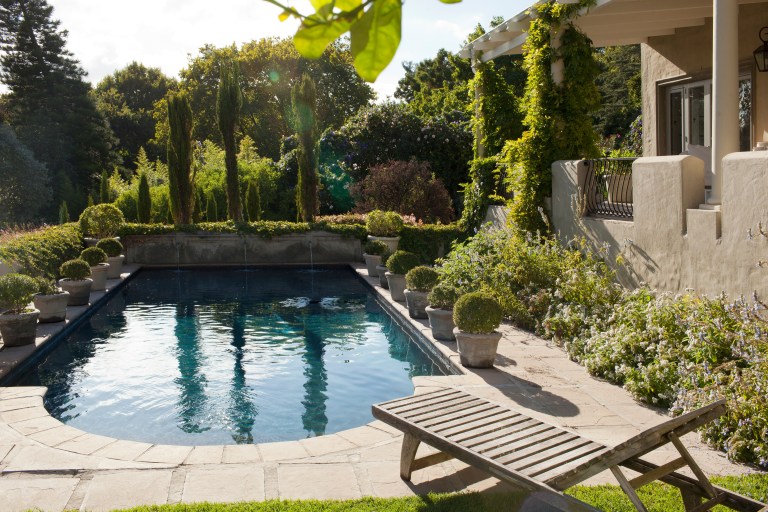 A private garden pool bordered by tall mature trees and ivy-covered walls that screen the space naturally, with a single wooden lounge chair at poolside.