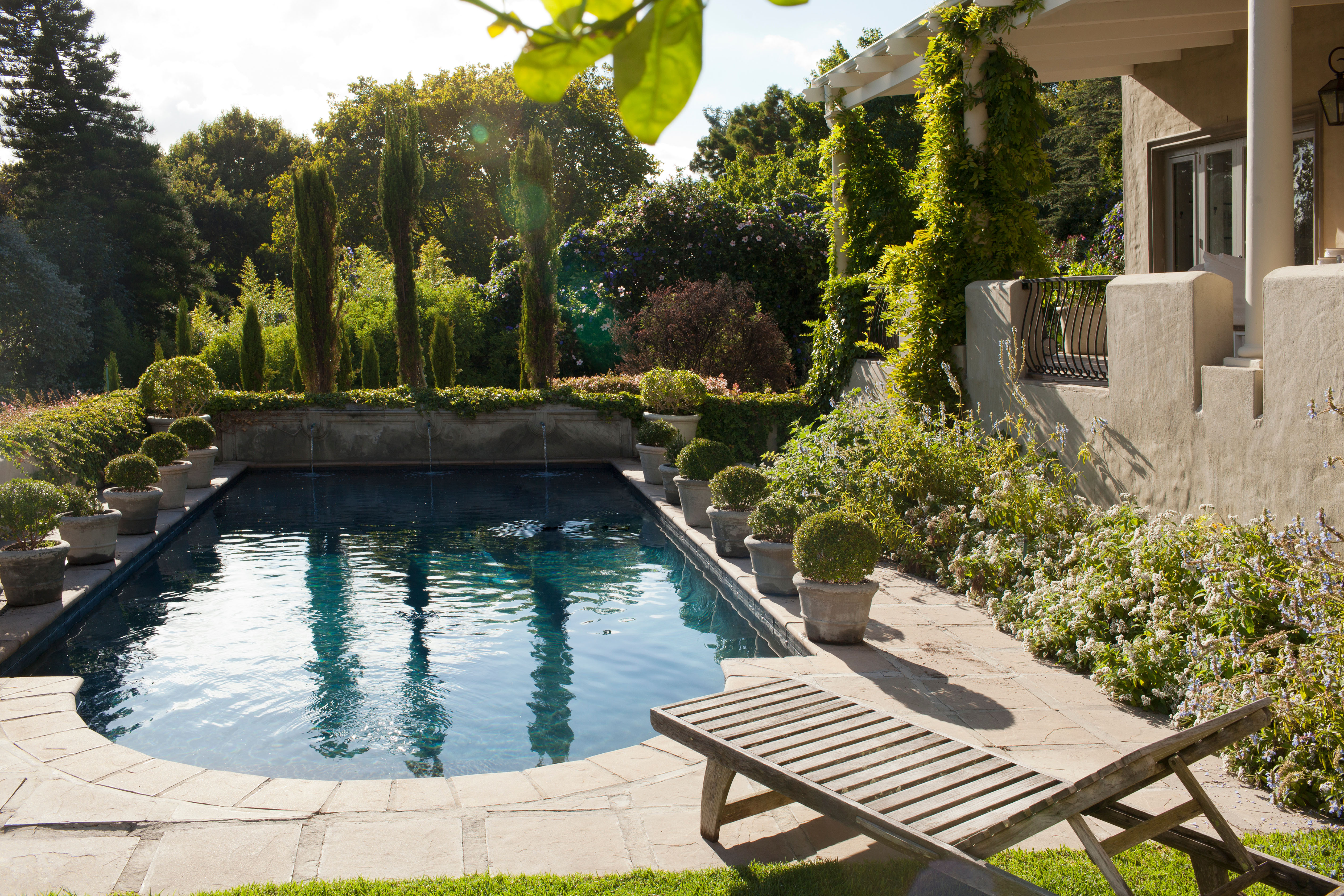 A private garden pool bordered by tall mature trees and ivy-covered walls that screen the space naturally, with a single wooden lounge chair at poolside.