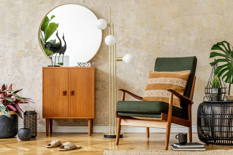 A mid-century wood cabinet anchoring a richly layered corner, with a round mirror, an emerald velvet armchair, and trailing greenery.