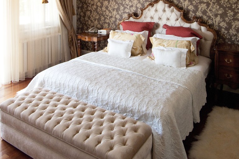 Dense antique-gold floral wallpaper covers the wall behind a carved cream headboard, dressed with a tufted bench and crystal chandelier light.