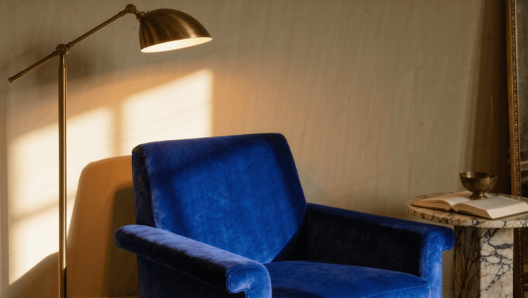 A sapphire velvet chair sits in a pool of warm lamp light while the rest of the room recedes into soft shadow.
