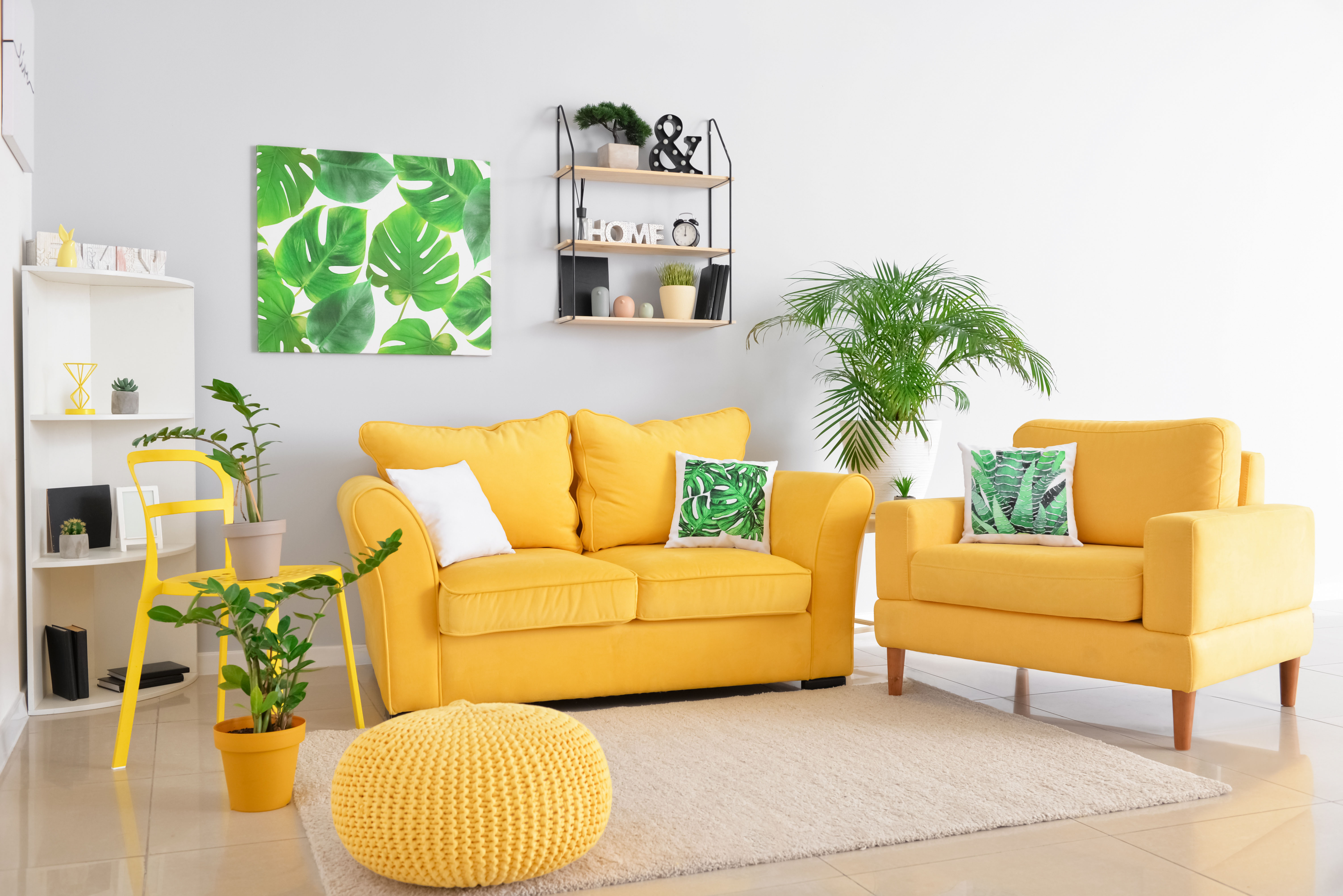 A mustard-yellow sofa and matching armchair face each other over a woven pouf and rug, layered with potted plants and botanical art on the wall.