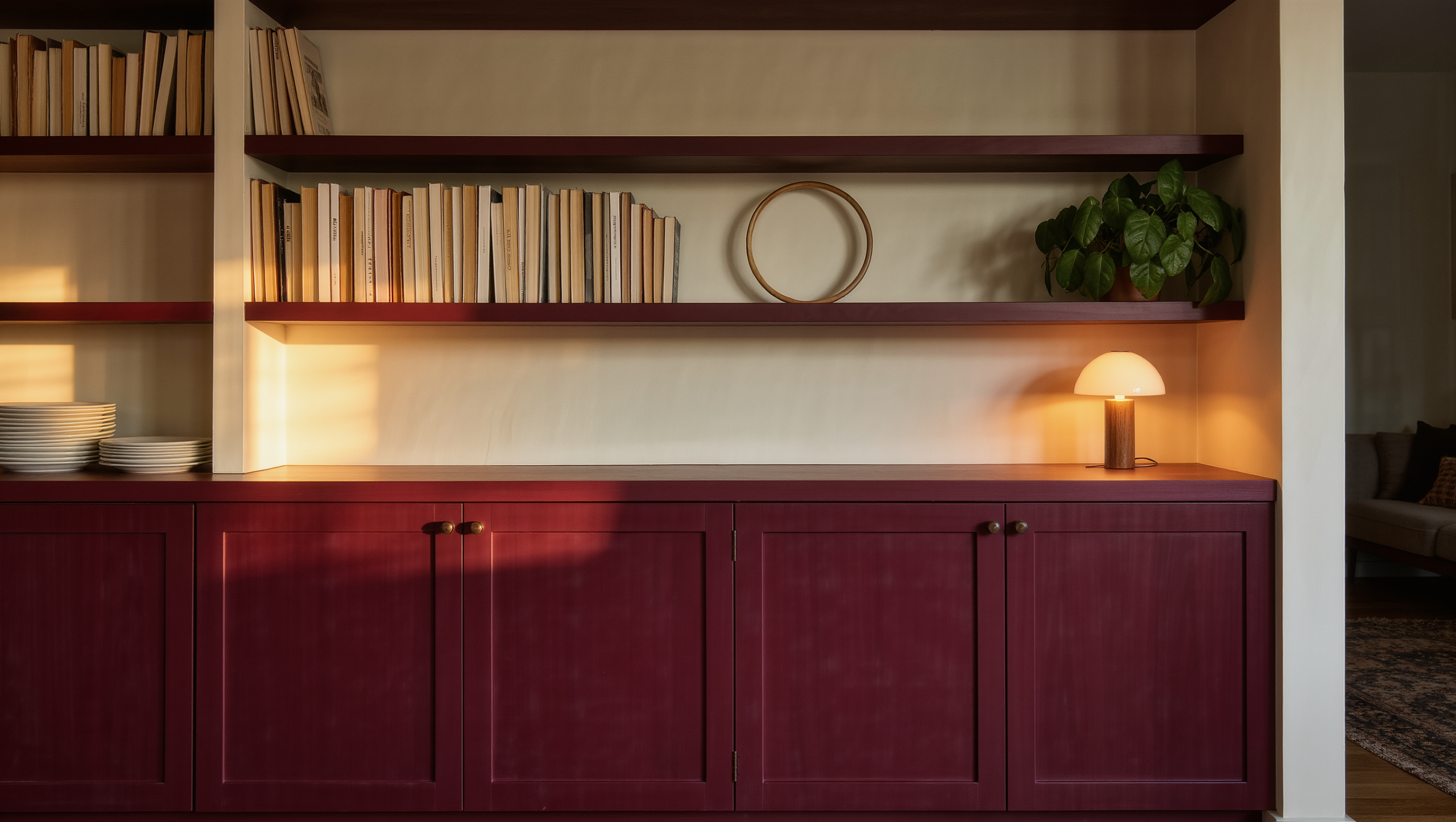 Storage wall pairing closed burgundy cupboards below with a few open shelves of daily objects above, one shelf left half empty