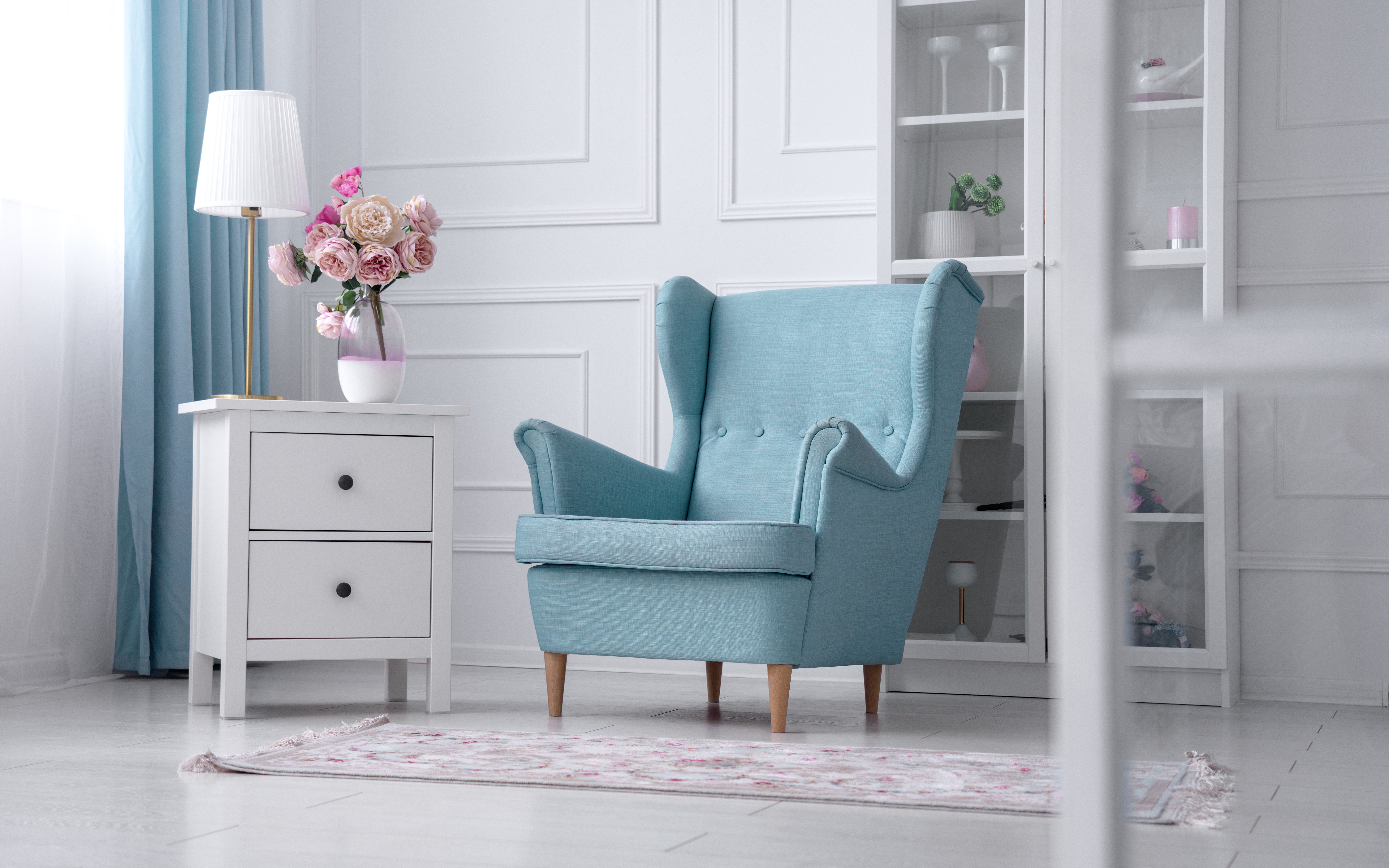 A sky-blue wingback armchair styled beside a white drawer cabinet topped with fresh flowers, set against a paneled wall with a curtained window.