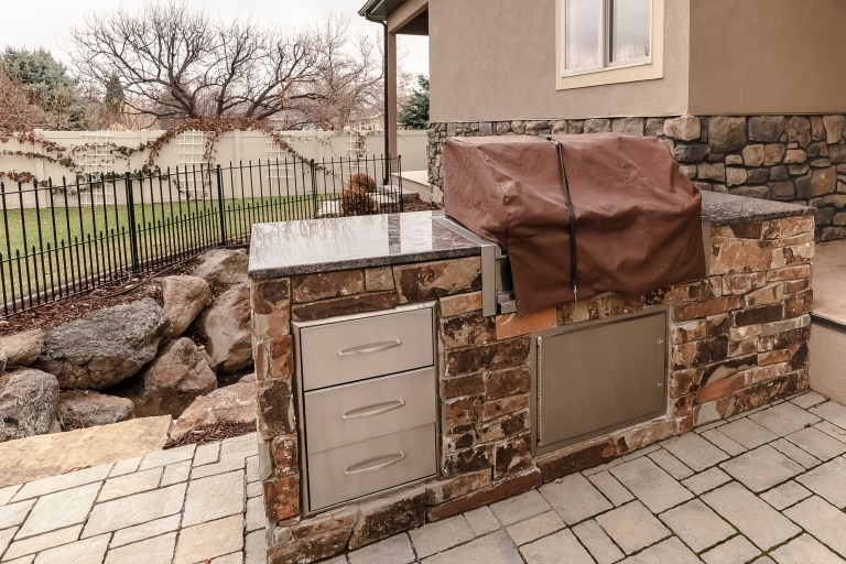 A stacked-stone outdoor kitchen counter with a built-in covered grill and stainless steel drawers, set on a paved patio.