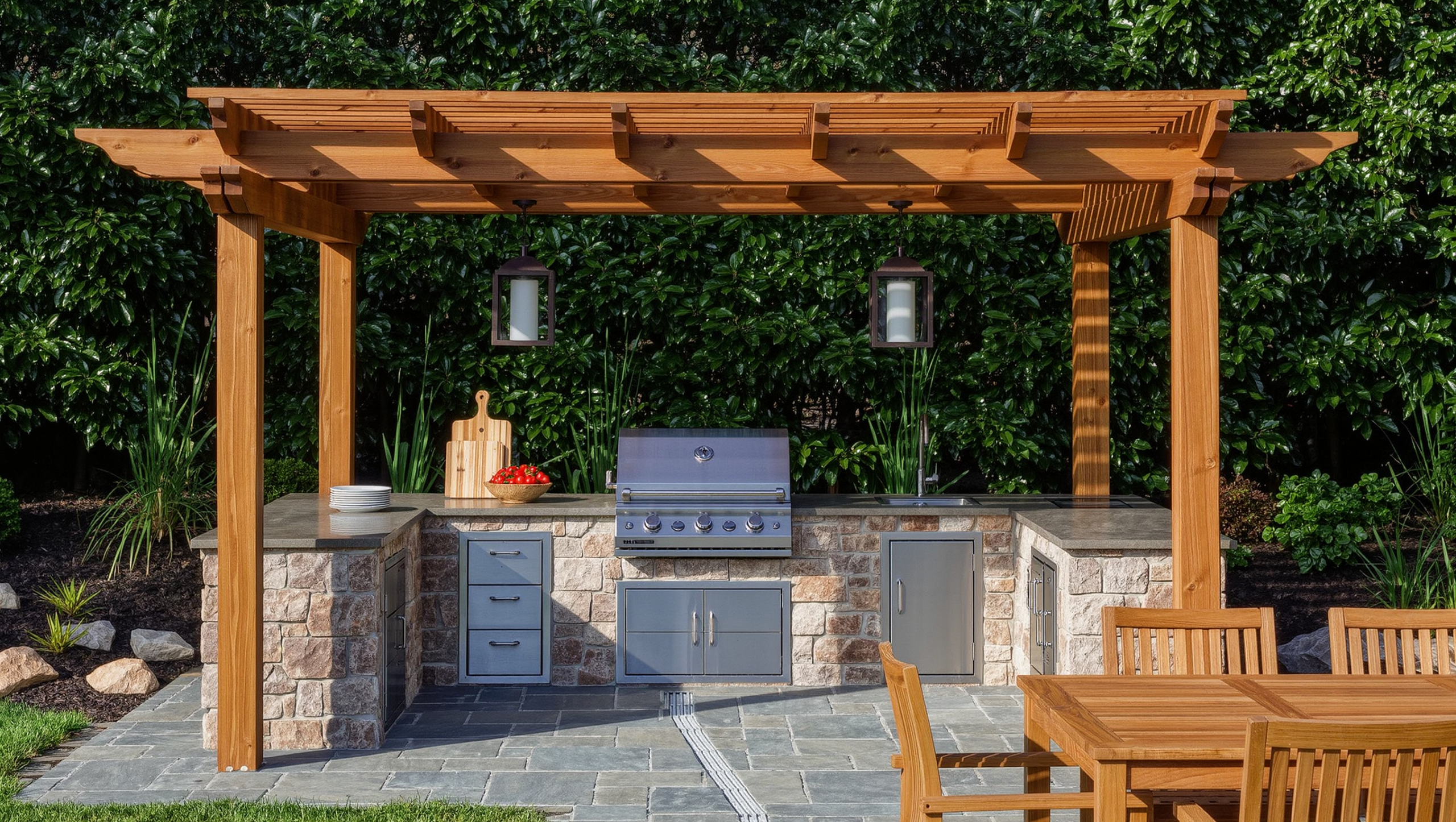 An outdoor kitchen with a grill set into a stone counter, generous landing space, storage drawers, and a shade pergola in late-morning sun.