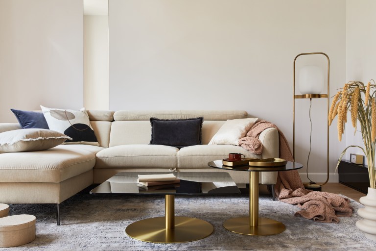 A deep sectional sofa in cream boucle spans the room, grounded by a textured rug and paired with two round brass coffee tables and a draped throw.