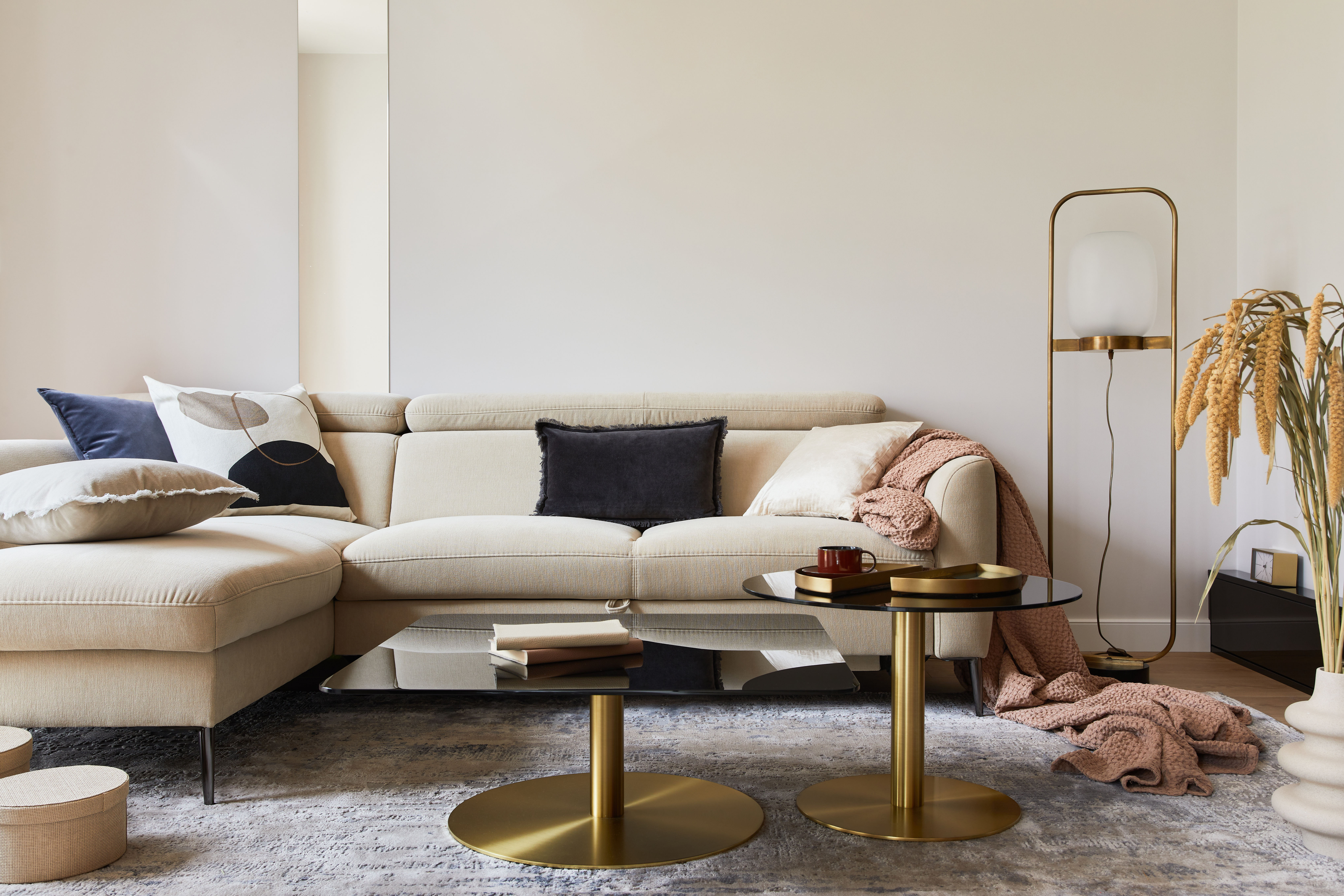 A deep sectional sofa in cream boucle spans the room, grounded by a textured rug and paired with two round brass coffee tables and a draped throw.
