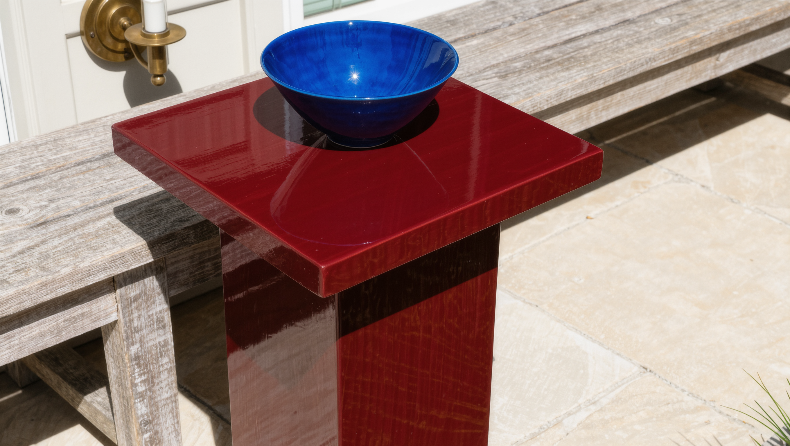 An oxblood lacquer outdoor side table beside a weathered teak bench on limestone