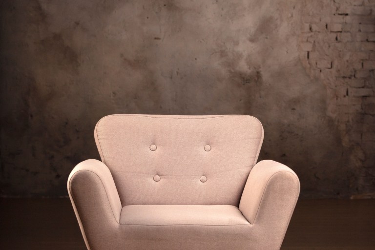A pale blush tufted armchair with dark wood legs stands alone against a moody charcoal textured wall.