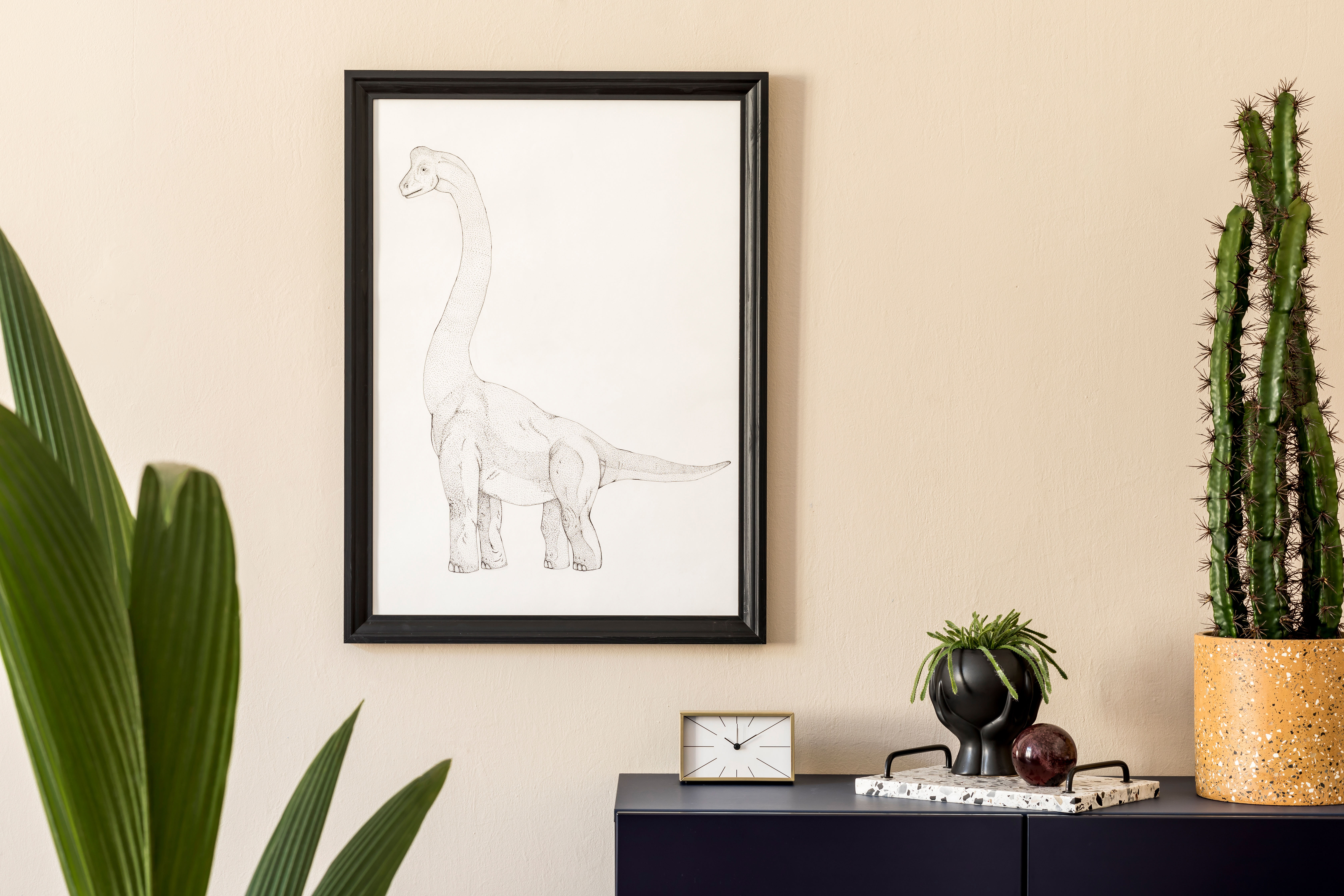 A dark navy-blue commode topped with a small gold clock and potted cactus sits beneath a framed pencil sketch on a warm beige wall.