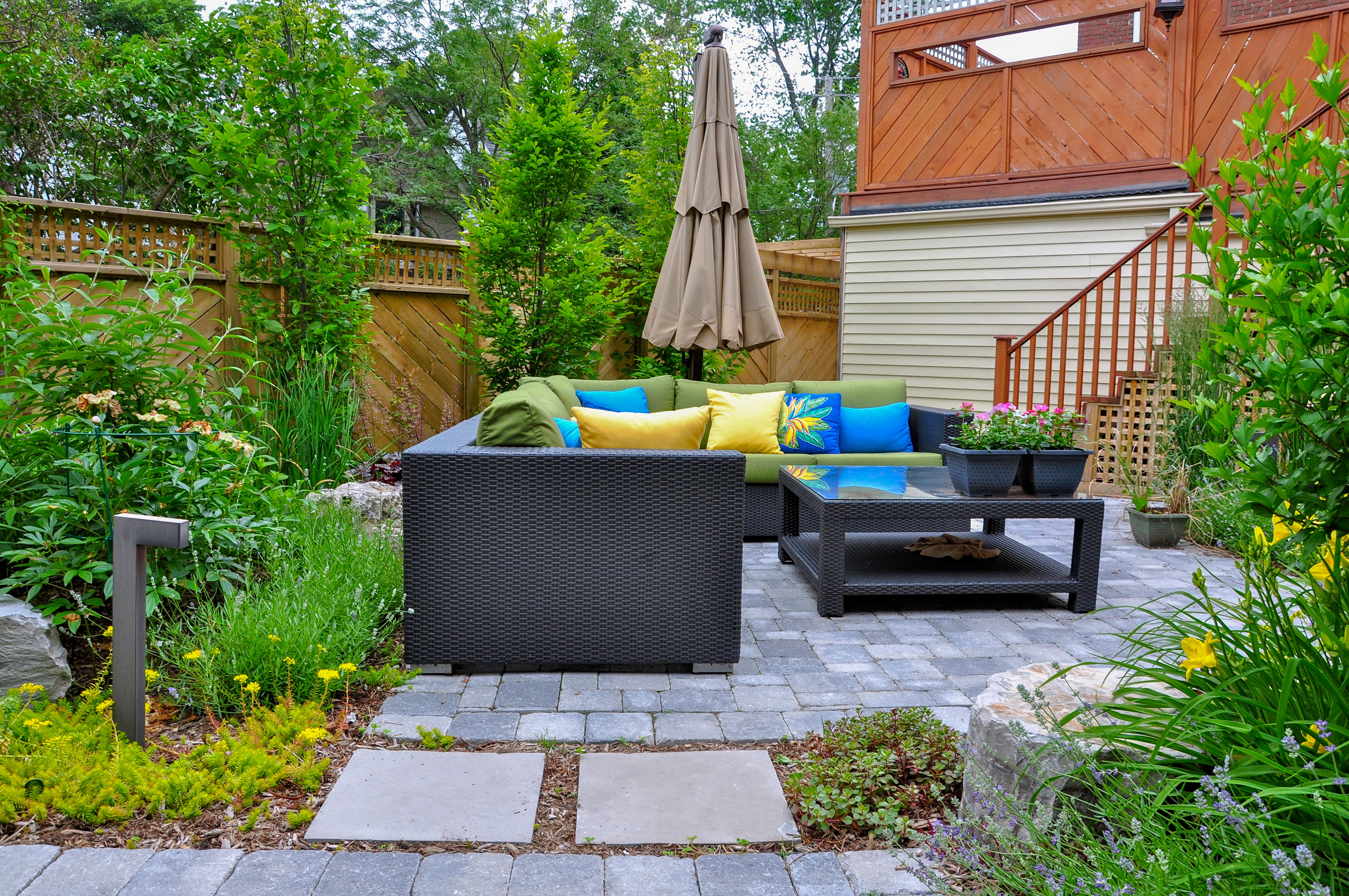 A paver-stone patio bordered by a boulder edge and dense plantings, with a modern outdoor sectional in the seating area.