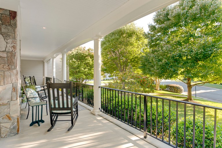 A stone-columned covered porch with black wrought-iron railing and a pair of dark wicker rocking chairs overlooking a leafy lawn.