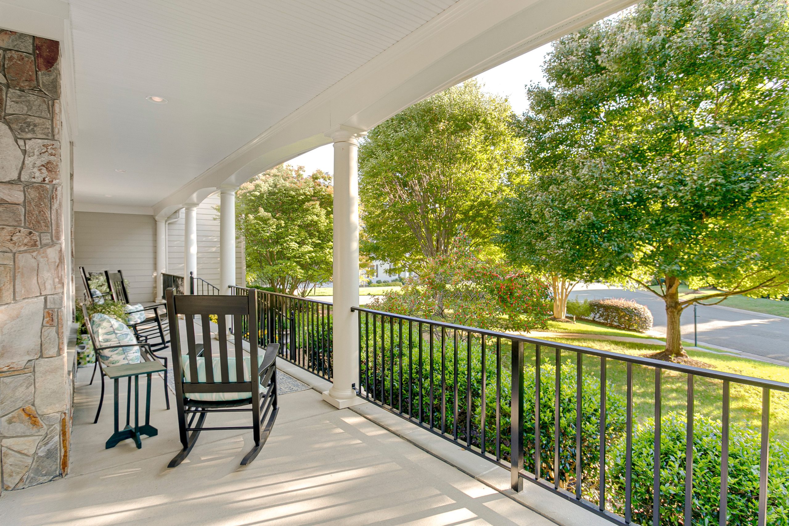 A stone-columned covered porch with black wrought-iron railing and a pair of dark wicker rocking chairs overlooking a leafy lawn.