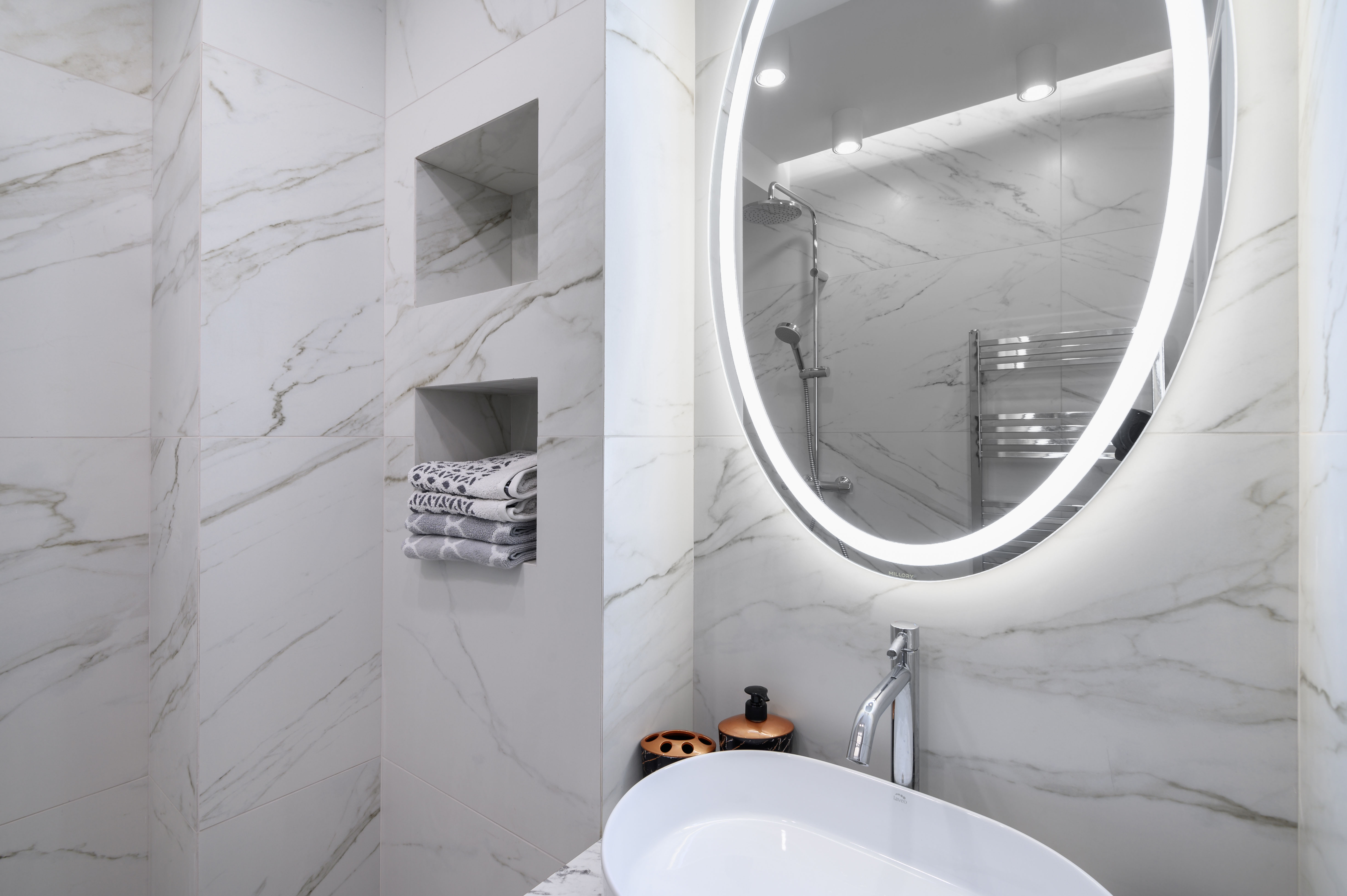 A marble-tiled bathroom with a backlit oval mirror above a freestanding oval sink, and recessed marble niche shelving built into the shower wall.