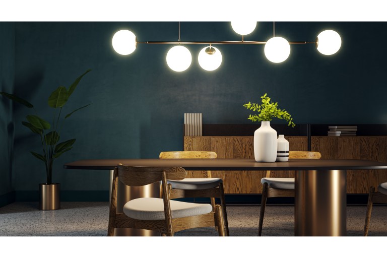 A wood-topped dining table on a brass pedestal base beneath a brass globe-cluster chandelier, set against a deep teal wall with a marble-topped console behind.