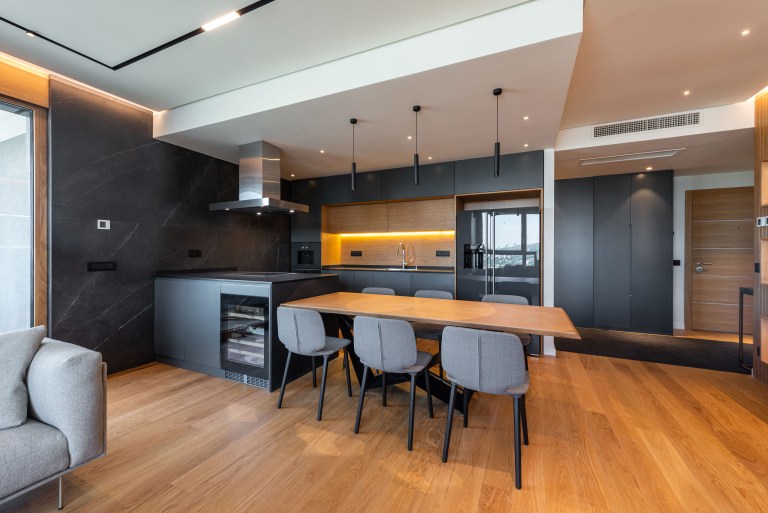 A polished walnut-toned dining table anchors a dark, modern kitchen with black cabinetry and brass pendant lighting.