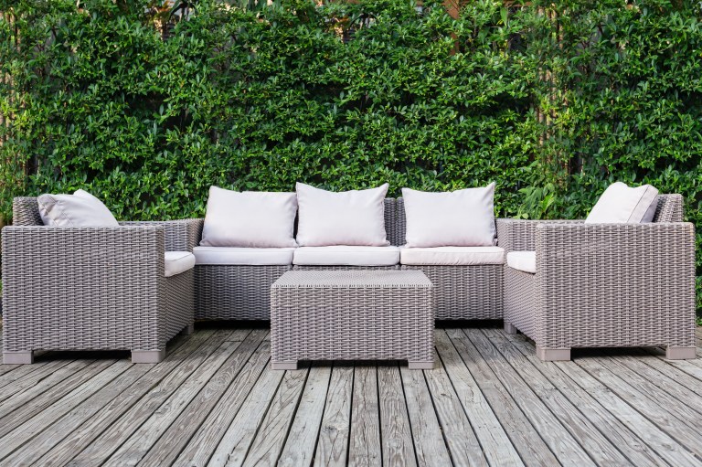 A beige rattan sectional sofa and ottoman are arranged on a weathered wood deck backed by a dense green hedge.