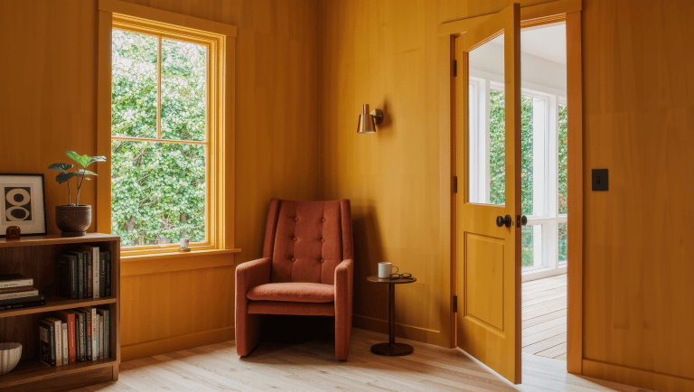 A bright ochre-toned room corner with a half-open door, its swing arc kept clear on pale oak flooring, beside a rust reading chair and bookshelf.