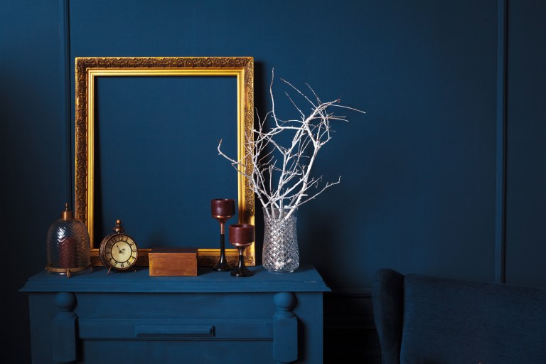 A deep sapphire-blue wall behind a matching console styled with a gilt-framed mirror, brass candlesticks, and the velvet arm of a chair at the frame's edge.