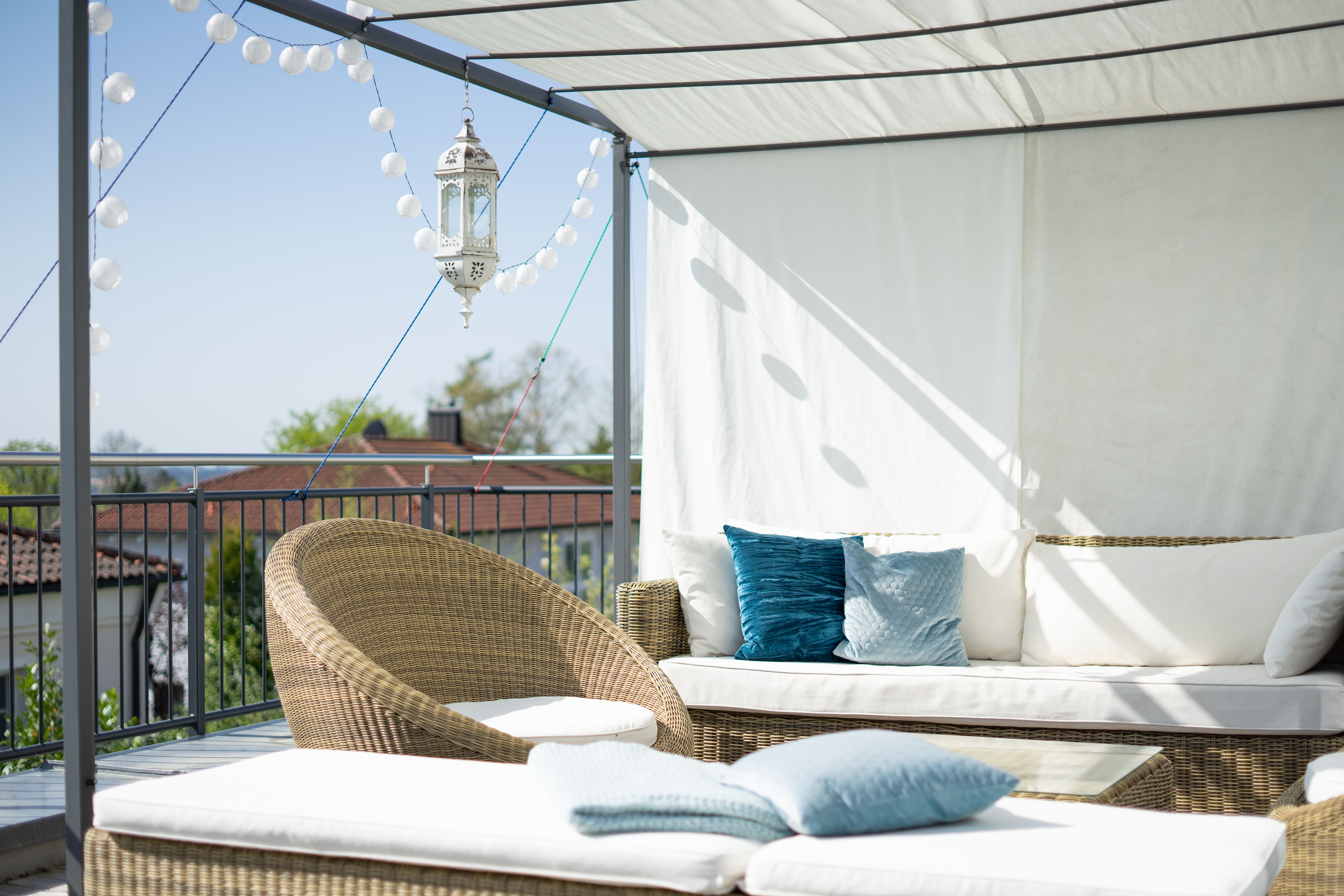 A rooftop terrace lounge with a fabric canopy overhead, wicker daybeds and chairs dressed in cream and teal cushions, string lights, and a hanging lantern.