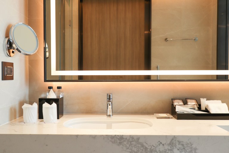 A backlit mirror glows above a veined marble vanity counter with a polished chrome faucet and neatly arranged bath amenities.