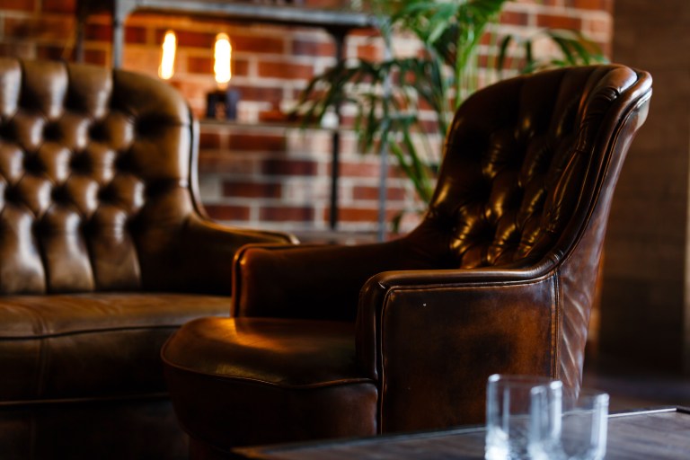 Tufted brown leather wingback chairs beside a glowing brick fireplace, styled with a potted plant for a warm library-like reading corner.