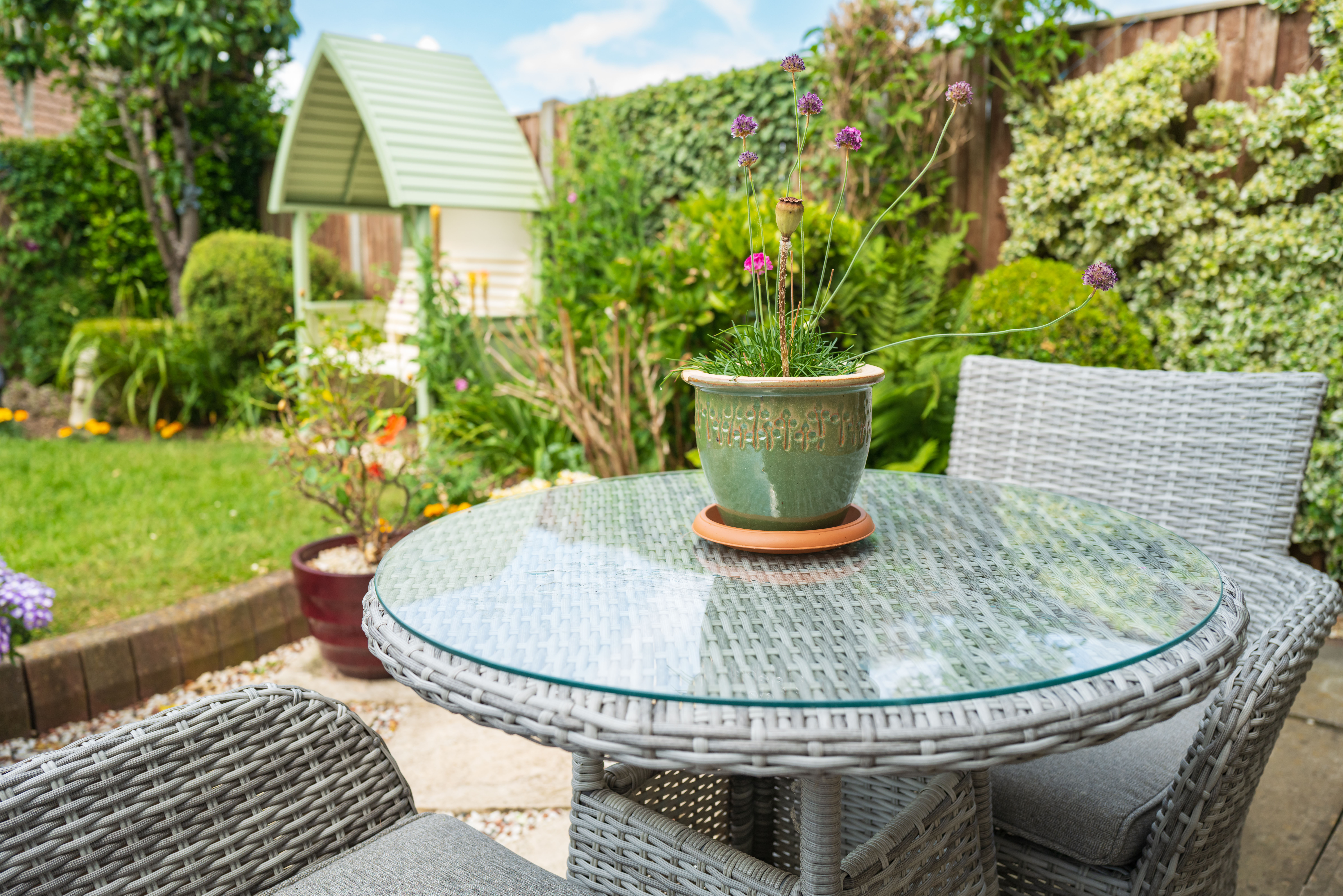 A round glass-top rattan table with matching chairs on a private garden patio, surrounded by lush greenery and a potted flowering plant.