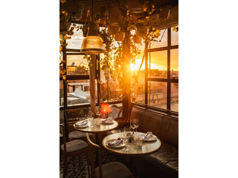 Two small round marble-top bistro tables set for wine on a glass-walled rooftop terrace, backlit by golden-hour sun through hanging greenery.