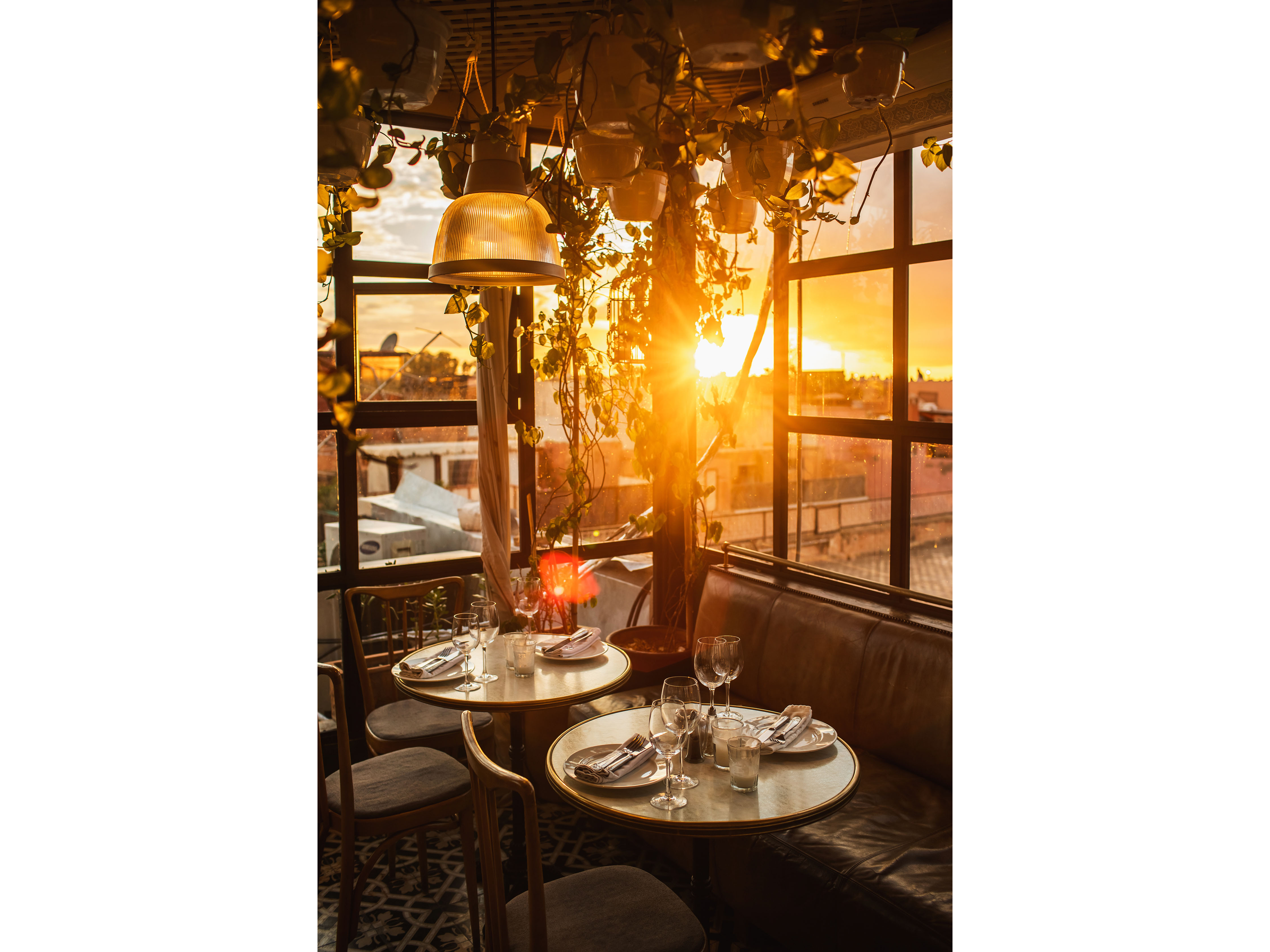 Two small round marble-top bistro tables set for wine on a glass-walled rooftop terrace, backlit by golden-hour sun through hanging greenery.