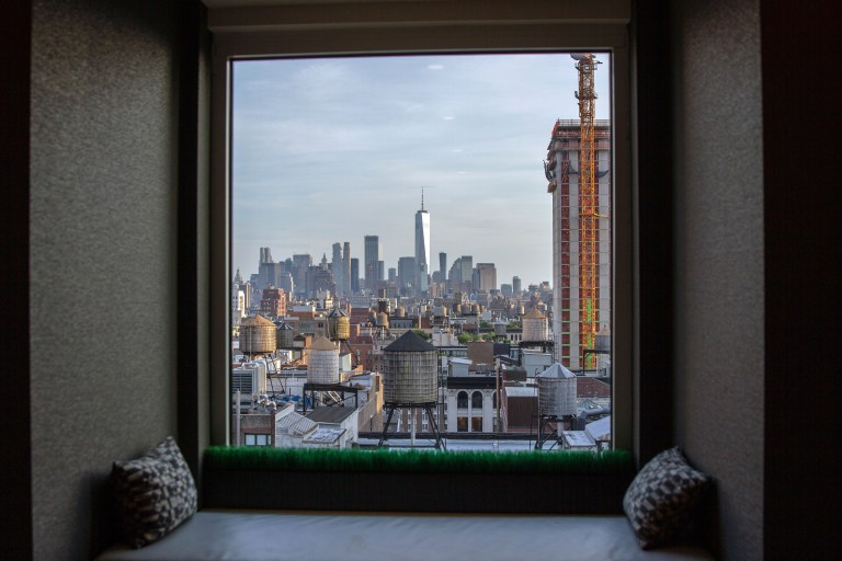 A cushioned window seat set into a deep bay window frames a sunlit view of a city skyline, natural light pouring across the nook's pillows.