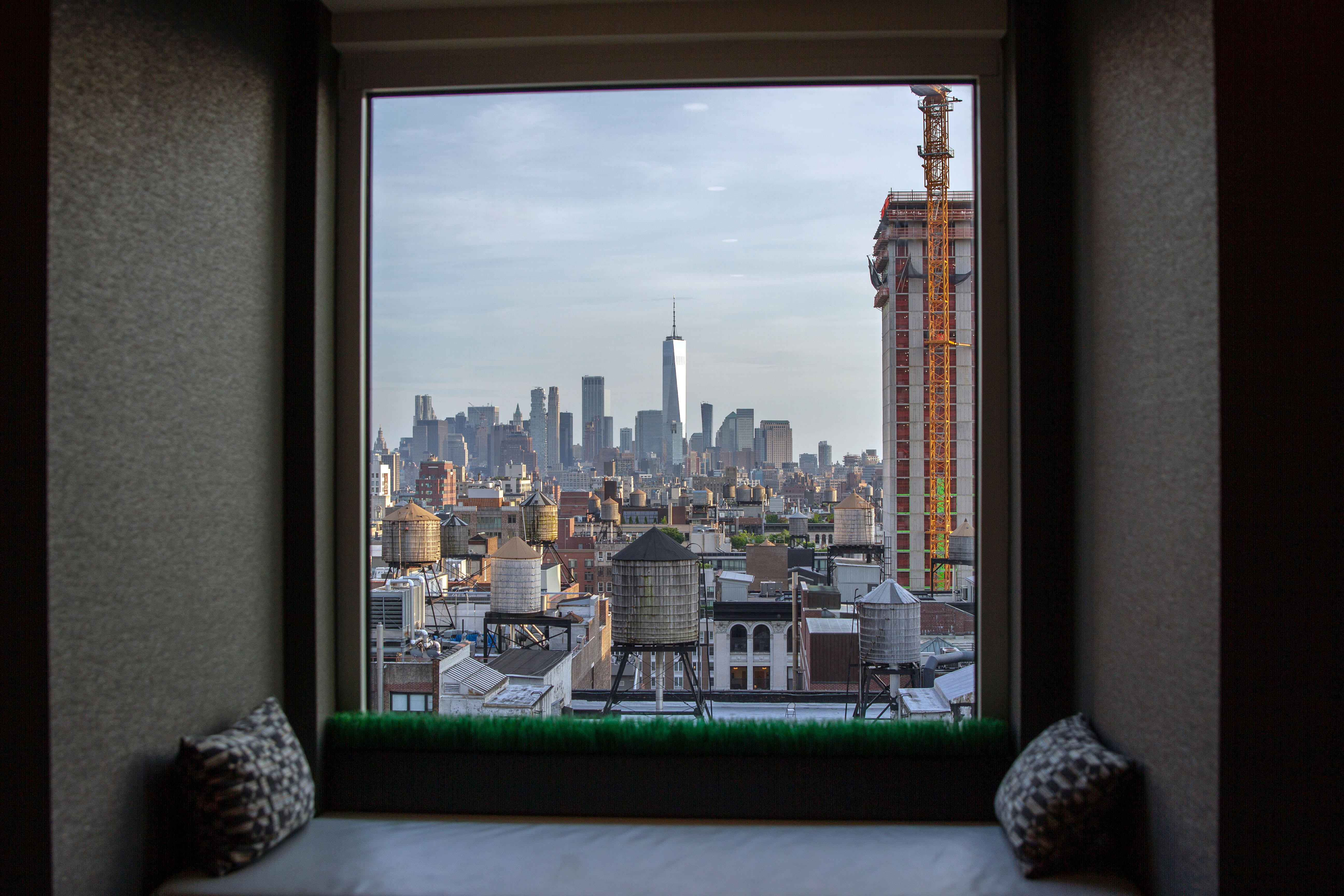 A cushioned window seat set into a deep bay window frames a sunlit view of a city skyline, natural light pouring across the nook's pillows.