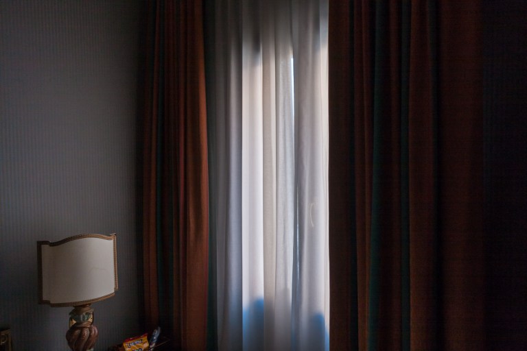 Deep rust and burgundy floor-to-ceiling curtains frame a glowing window in a moody bedroom, with a vintage lamp glimpsed at the edge of frame.