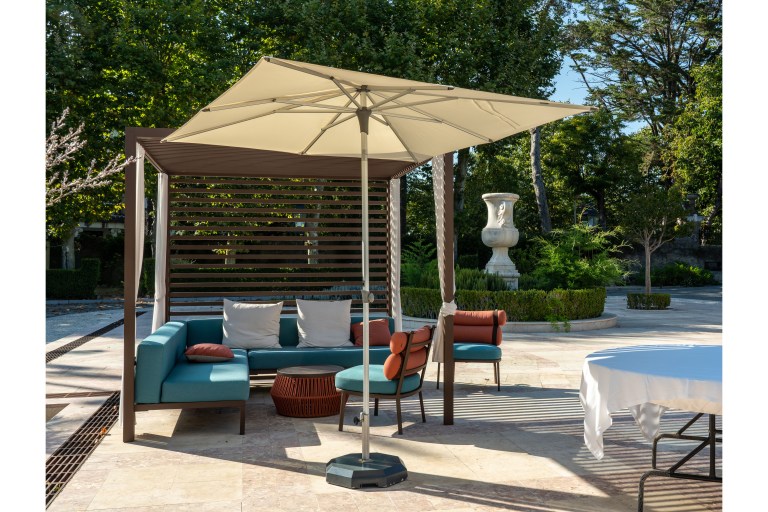 A marble-paved terrace corner shaded by a cantilevered umbrella, furnished with teal lounge seating, rust-orange pillows, and a classical urn in the garden beyond.