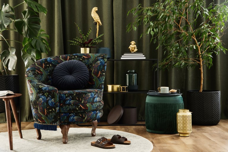 A richly layered corner with deep green velvet drapery, a patterned jewel-toned armchair, brass accent tables, and a grouping of tall potted plants against dark walls.